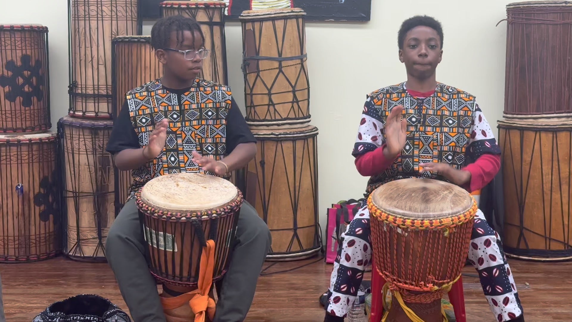Two students clapping and playing djembe together in front of a wall of drums