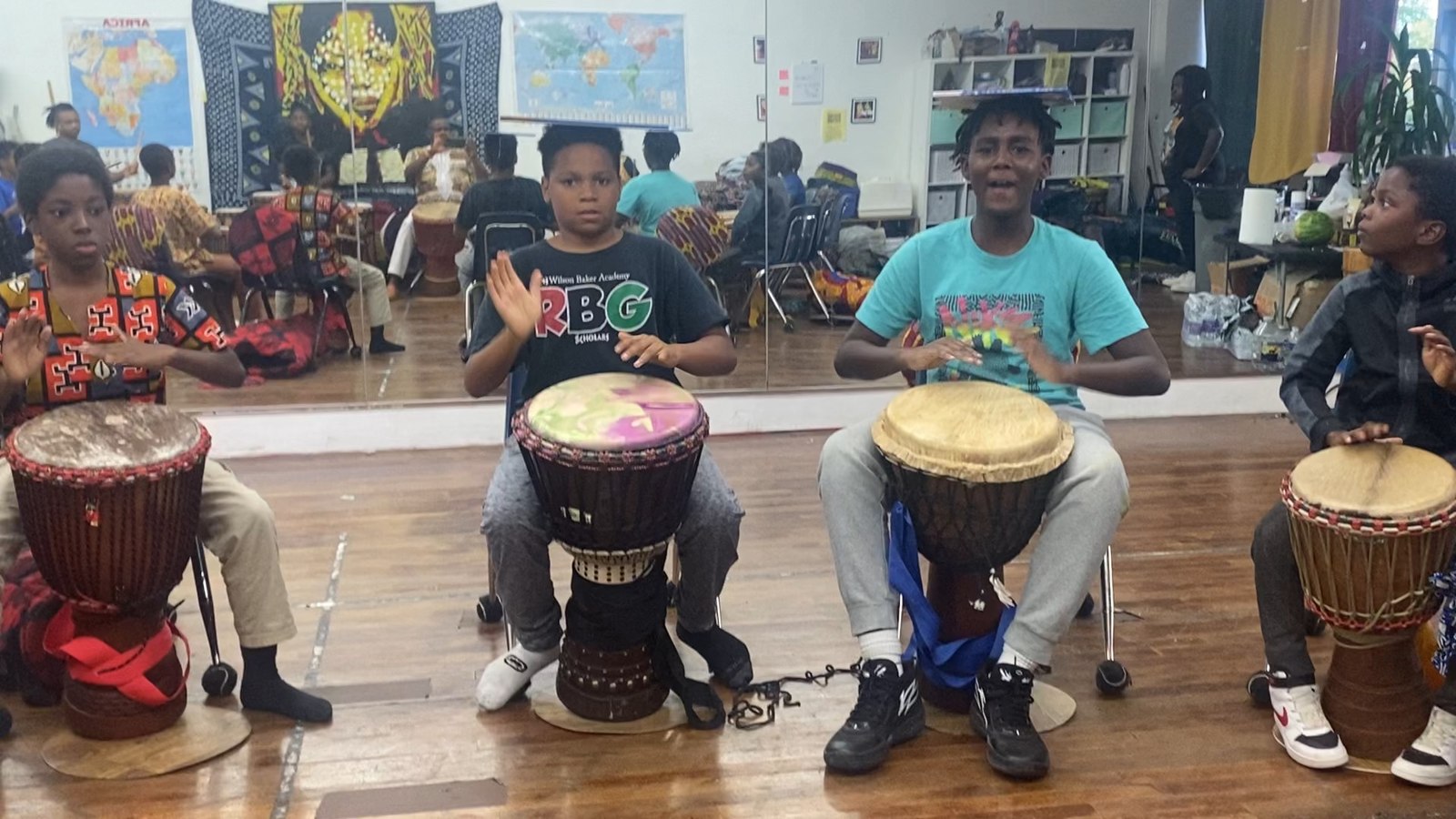 Two teen students clapping and drumming together in the studio, African maps on the wall behind them