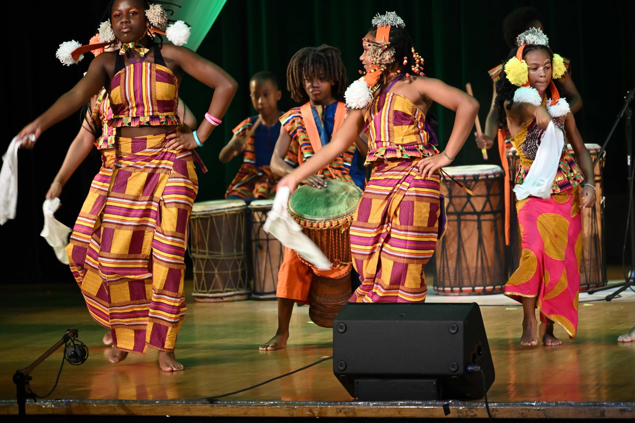 Young dancers in kente-patterned outfits and pom-pom headpieces performing at Birthright, drummers behind them