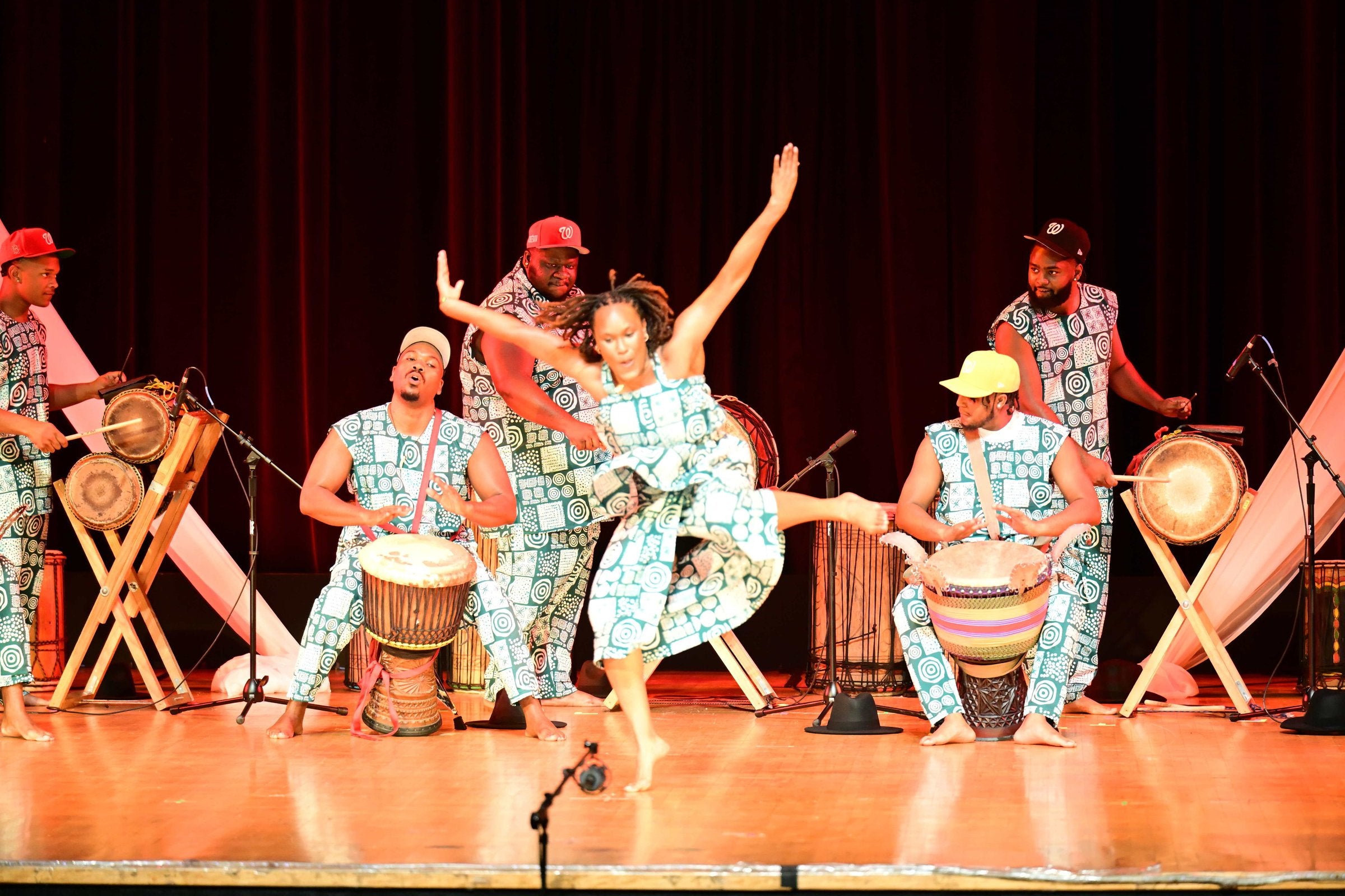 A dancer leaping mid-air at center stage, surrounded by six drummers in matching patterned outfits at Birthright