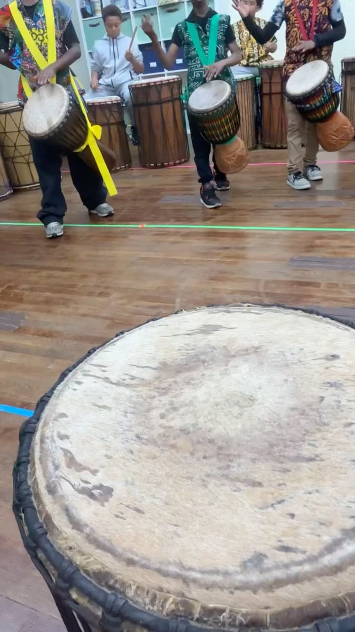 Close-up of a djembe skin and rope work, with students drumming in the background