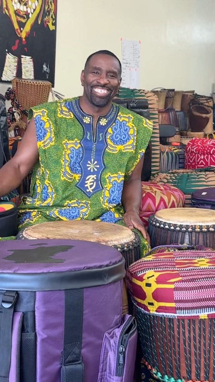 Mahiri Fadjimba Keita smiling behind a row of djembes in the studio
